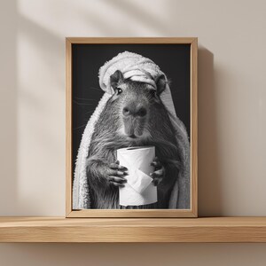 May include: Monochrome framed print of a capybara, wrapped in a towel, clutching a roll of toilet paper. The artwork is displayed on a wooden shelf. The image has a humorous, lighthearted feel, with the capybara as the main subject.