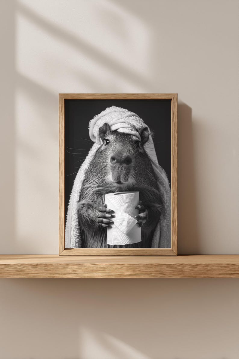 May include: Monochrome framed print of a capybara, wrapped in a towel, clutching a roll of toilet paper. The artwork is displayed on a wooden shelf. The image has a humorous, lighthearted feel, with the capybara as the main subject.