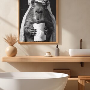 May include: Monochrome framed print featuring a capybara wrapped in a towel, clutching a toilet roll. The image is set in a bathroom with a wooden shelf, a white sink, and a bathtub.
