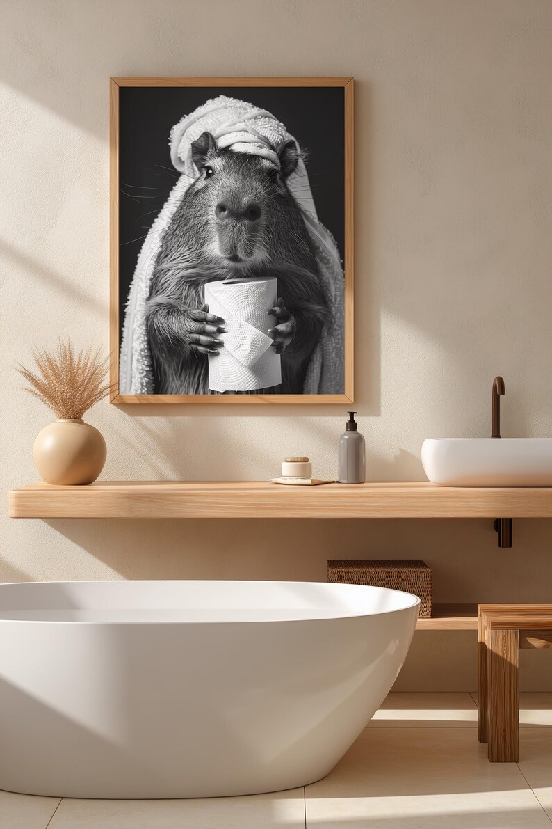 May include: Monochrome framed print featuring a capybara wrapped in a towel, clutching a toilet roll. The image is set in a bathroom with a wooden shelf, a white sink, and a bathtub.