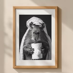 May include: Monochrome framed print of a capybara in a towel, clutching a toilet roll. The image has a humorous theme, suitable for bathroom decor or as a quirky gift. The print is framed in a wooden frame.