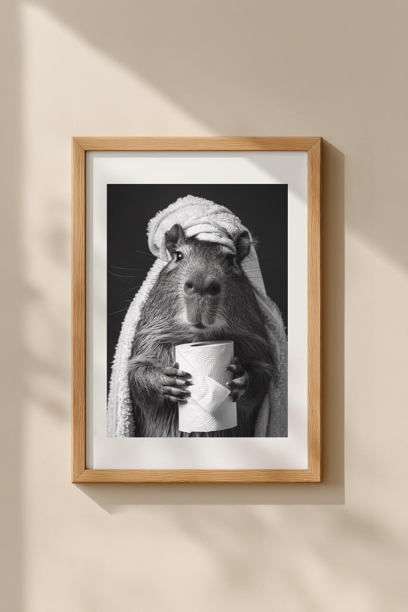 May include: Monochrome framed print of a capybara in a towel, clutching a toilet roll. The image has a humorous theme, suitable for bathroom decor or as a quirky gift. The print is framed in a wooden frame.
