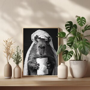 May include: Monochrome framed print showing a capybara in a towel, clutching a toilet roll. The artwork sits on a wooden shelf with vases and a houseplant. The image has a humorous theme and is a fun piece of wall art.