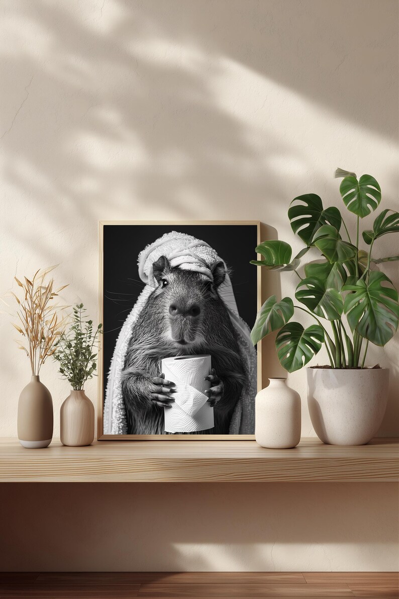May include: Monochrome framed print showing a capybara in a towel, clutching a toilet roll. The artwork sits on a wooden shelf with vases and a houseplant. The image has a humorous theme and is a fun piece of wall art.