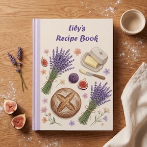 Könnte beinhalten: Ein gebundenes Rezeptbuch mit dem Titel Lily's Recipe Book und einem cremefarbenen Einband, der mit Lavendelsträußen, einem Laib Brot, Feigen, einer Butterdose und einem Buttermesser illustriert ist. Das Buch hat einen hellvioletten Buchrücken.