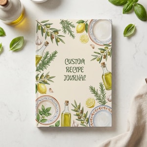 Könnte beinhalten: Ein rechteckiges Rezeptbuch mit cremefarbenem Einband und dem Text CUSTOM RECIPE JOURNAL in Dunkelgrün. Der Einband ist mit Illustrationen von Olivenzweigen, Zitronen, Knoblauchzehen, Rosmarinzweigen, Ölflaschen aus Glas und Tellern verziert.