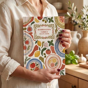 May include: A hardback recipe book with a white cover featuring a colourful pattern of plates, lemons, tomatoes, figs, garlic, and herbs. The central text reads Harrington Family Recipe Book in a decorative frame with green and red leaf accents.