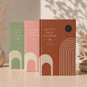 Pode incluir: Três agendas diárias em verde, rosa e terracota. Cada capa apresenta o texto "KATIE'S DAILY PLANNER" e "SLOW DOWN STAY FOCUSED" com um gráfico de um arco cor de creme e um círculo escuro na capa da agenda na cor terracota.