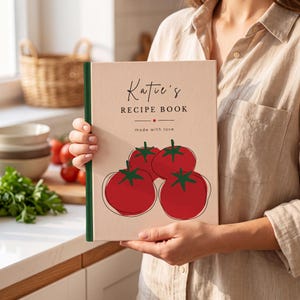 May include: A hardback recipe book with a light beige cover featuring a graphic of four red tomatoes with green stems. The text on the cover reads Katie's RECIPE BOOK made with love. The book has a dark green spine and a rectangular shape.