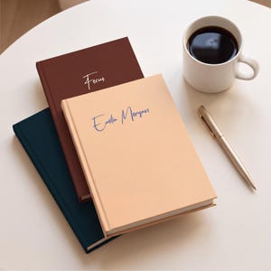 May include: Three hardback notebooks stacked together in peach, dark brown, and dark blue. The peach notebook features the name Emilia Morgan in blue script, while the brown notebook has the word Focus in white script. A gold pen and a white mug of coffee.