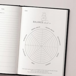 May include: Open journal with a black cover, displaying a "Balance Check-in" chart. The chart is a circular diagram divided into sections for mental health, physical health, relationships, career, finances, home, and spirituality.