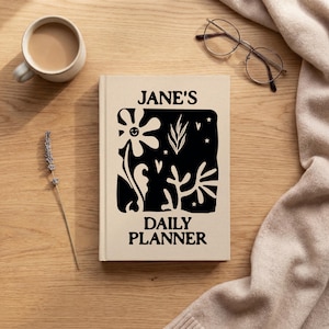 May include: A beige daily planner with the text "JANE'S DAILY PLANNER" and a black floral design. The planner is on a wooden surface, with a cup of coffee, glasses, and a sprig of lavender nearby.