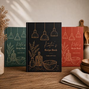 Puede incluir: Tres libros de recetas de tapa dura en negro, verde oscuro y rojo terracota. Cada libro presenta ilustraciones de líneas doradas de lámparas colgantes, un jarrón con ramas, una tabla de cortar y un cuenco, con el texto Katie's Recipe Book made with love.