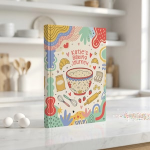 Op de afbeelding: Een hardcover boek getiteld Katie's Baking Journey met een crèmekleurige kaft voorzien van kleurrijke illustraties van een mengkom, garde, deegroller, meelzak, eieren, aardbeien en croissants omringd door abstracte vormen.