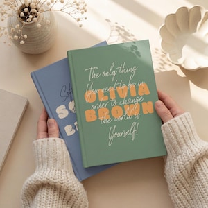 May include: Two hardcover journals, one sage green and one dusty blue. The green journal features the text 'The only thing you need to be in order to change the world is Yourself!' with the name 'OLIVIA BROWN' in large orange letters.