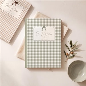 Puede incluir: Un libro de recetas de tapa dura con estampado de cuadros vichy en verde salvia y blanco, con una etiqueta central blanca que muestra un icono de lazo y el texto "Our Family Recipes" y "made with love". Se ve otro libro vichy beige.