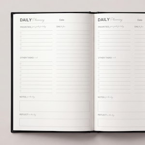 May include: Open black notebook with the words "DAILY Planning" at the top of each page. The pages are lined with sections for priorities, tasks, notes, and reflection. The notebook is open, displaying two facing pages.