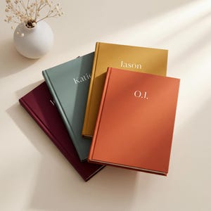 Peut inclure: Quatre carnets à couverture rigide en bordeaux, vert sauge, jaune moutarde et orange brûlé. Chaque carnet présente un nom ou des initiales personnalisés imprimés en blanc sur la couverture. Les carnets sont disposés en éventail.
