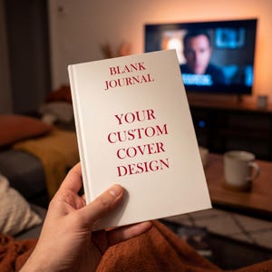 May include: A white blank journal with the text "BLANK JOURNAL" and "YOUR CUSTOM COVER DESIGN" in red. The journal is held in a person's hand, with a blurred background featuring a television and a coffee mug.