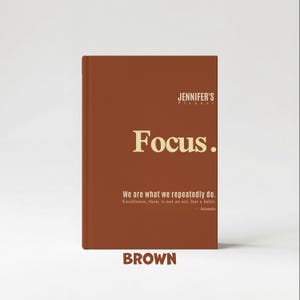 May include: A brown planner with the text "Focus." in gold lettering. The planner also features the text "We are what we repeatedly do. Excellence, then, is not an act, but a habit. - Aristotle" and the word "BROWN".