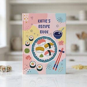 May include: A recipe book with a pink cover, featuring the text "Katie's Recipe Book." The cover design includes sushi illustrations, a plate of sushi, chopsticks, and abstract shapes. The book is on a white surface.