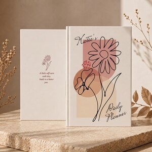 May include: A cream-coloured daily planner featuring a black line-art drawing of two flowers over muted pink and tan geometric shapes. The cover text reads Katie's Daily Planner. The back cover displays a small flower icon and the text A little self-care each day, leads to a better you.