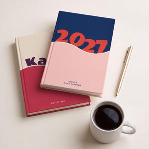 Könnte beinhalten: Zwei Hardcover-Planer mit wellenförmigem Farbblock-Design. Der obere Planer ist marineblau und hellrosa mit dem Text 2027, Katie's Daily Planner. Der untere Planer ist cremefarben und dunkelrot mit dem Text Ka und Day by Day. Ein goldener Stift und eine weiße Tasse mit schwarzem Kaffee liegen daneben.