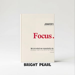 May include: A cream-colored planner with the text "JENNIFER'S Planner" at the top and the word "Focus." in red. Below, it reads "We are what we repeatedly do." and "BRIGHT PEARL" at the bottom.