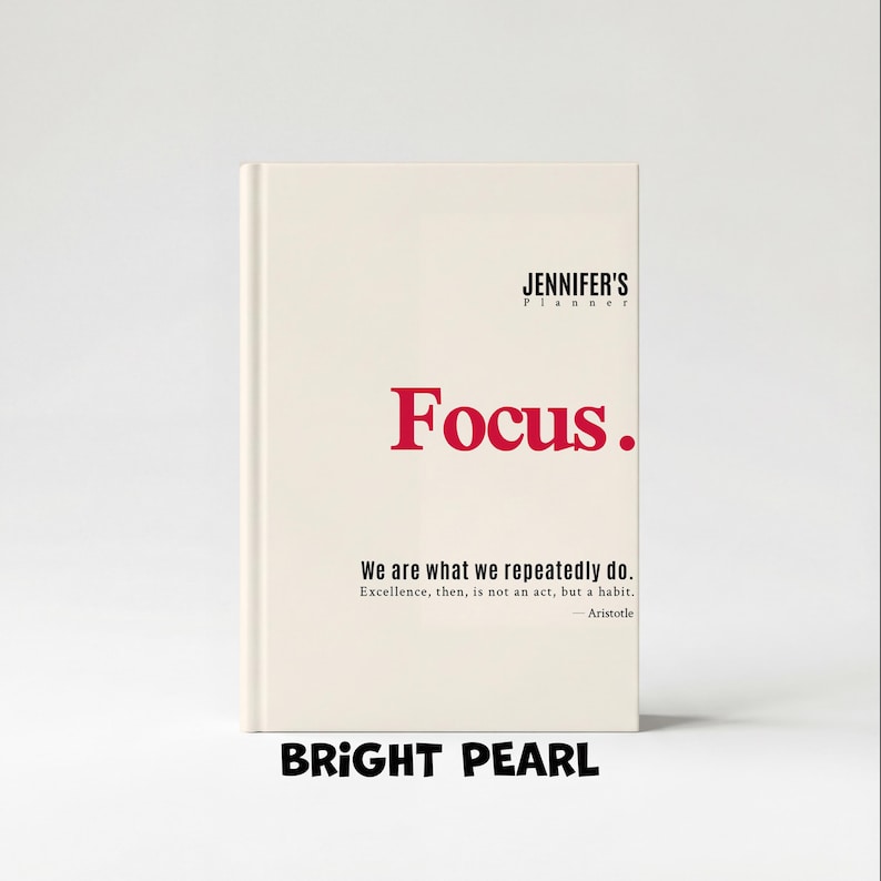 May include: A cream-colored planner with the text "JENNIFER'S Planner" at the top and the word "Focus." in red. Below, it reads "We are what we repeatedly do." and "BRIGHT PEARL" at the bottom.