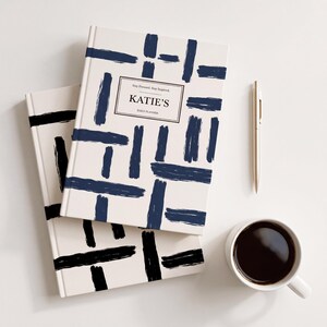 May include: Two hardback planners with a cream background and dark blue brushstroke grid pattern. The top planner features a rectangular label with the text "Stay Focused. Stay Inspired. KATIE'S DAILY PLANNER." A gold pen and a white mug of coffee.