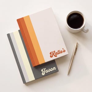 May include: Two rectangular notebooks with vertical stripes. The top notebook is cream with orange and rust stripes and the text Katie's. The bottom notebook is dark grey with light grey and cream stripes and the text Jason. A gold pen and coffee mug.