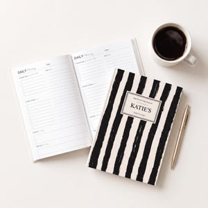 May include: A daily planner with a black and white vertical brushstroke pattern, featuring a central label that reads "Stay Focused. Stay Inspired. KATIE'S DAILY PLANNER." The planner is shown open to reveal lined pages for priorities and tasks.