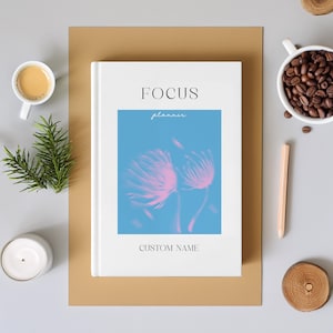 May include: A white planner with the word "FOCUS" at the top and "planner" in cursive below it. A blue rectangle with pink floral art is in the center. The words "CUSTOM NAME" are at the bottom. The planner is on a brown surface.