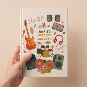以下が含まれることがあります： 音楽をテーマにしたクリーム色のジャーナル。表紙には、ギター、マイク、ヘッドフォン、スピーカー、ドラムセット、カセットテープのイラストが描かれています。「JACKIE'S CONCERT JOURNAL」の文字が中央に表示されています。
