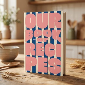Puede incluir: Un libro de recetas de tapa dura con rayas verticales azules y crema. La portada presenta un texto grande, grueso y de color rosa sin remates que dice OUR BOOK OF RECIPES. El libro se mantiene en posición vertical.