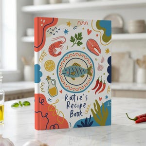 Op de afbeelding: Een receptenboek met harde kaft en een crèmekleurige omslag met kleurrijke illustraties van een vis op een bord, garnalen, zalm, chilipepers, knoflook, citroen en olijfolie. De tekst luidt Katie's Recipe Book in donkerblauwe letters.
