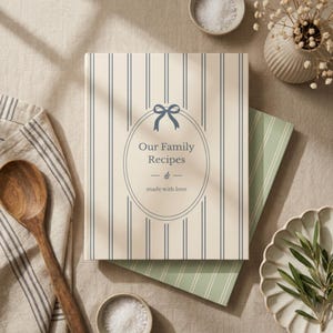 May include: A hardcover cookbook titled "Our Family Recipes" with a cream-colored cover featuring vertical blue stripes and an oval design. The book rests on a green striped surface, with a wooden spoon and a small bowl of salt nearby. A small vase with white flowers is also visible.