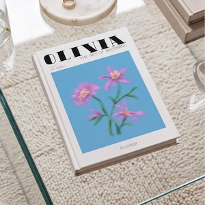 May include: A white planner with the text OLIVIA THE ART OF BECOMING, VOL 1, 2027 EDITION, and PLANNER printed in black. The cover features a central blue rectangular graphic containing a soft-focus illustration of three pink flowers.