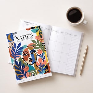 以下が含まれることがあります： カラフルな抽象的な顔のイラスト、花柄のモチーフ、そして「KATIE'S DAILY PLANNER」と「dream | plan | do」という文字が表紙に描かれたデイリープランナー。プランナーは月間カレンダーのページが開かれており、コーヒーカップとゴールドのペンが隣に置かれています。