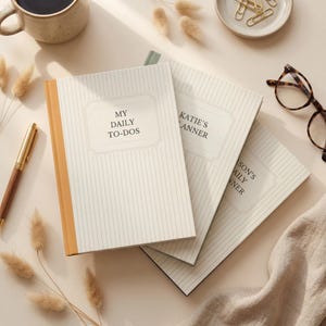 May include: Three notebooks with cream-colored covers and vertical pinstripes are displayed on a light beige surface. The first notebook reads "MY DAILY TO-DOS" and has a tan spine. The other two notebooks read "KATIE'S PLANNER" and "SON'S DAILY PLANNER".
