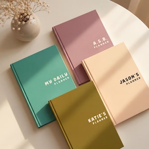 May include: Four rectangular planners with solid-coloured covers in dusty rose, teal, light peach, and olive green. The teal planner reads MY DAILY PLANNER, the rose one reads A.S.B. PLANNER, the peach one reads JASON'S PLANNER, and the olive one reads KATIE'S PLANNER.