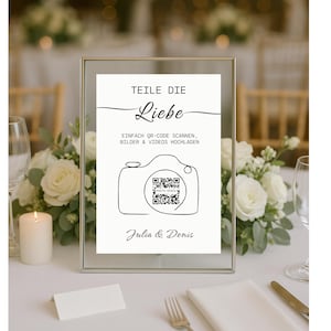 May include: A framed sign with the text "TEILE DIE Liebe" and instructions to scan a QR code to upload photos and videos. The sign features a line drawing of a camera and is signed "Julia & Denis". The sign is on a table with flowers.