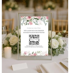 Könnte beinhalten: Ein gerahmtes Hochzeitsschild mit floralen Akzenten und Text auf Deutsch. Das Schild lädt die Gäste ein, über einen QR-Code einem Fotoalbum beizutreten. Das Design zeigt rosa Rosen und Grün, auf einem Tisch mit Kerzen und Blumen.