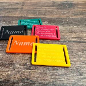 Könnte beinhalten: Eine Sammlung farbiger, rechteckiger Namensschilder in Schwarz, Orange, Rot, Grün und Gelb. Jedes Schild hat einen Schlitz und gravierten Text, mit dem Wort "Name" oder Kontaktinformationen.