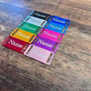 Könnte beinhalten: Eine Sammlung farbiger rechteckiger Anhänger, auf denen jeweils das Wort "Name" in Weiß aufgedruckt ist. Die Anhänger sind in verschiedenen Farben erhältlich, darunter Schwarz, Silber, Grün, Gelb, Rot, Blau, Pink, Lila und Hellrosa. Sie liegen auf einer Holzoberfläche.