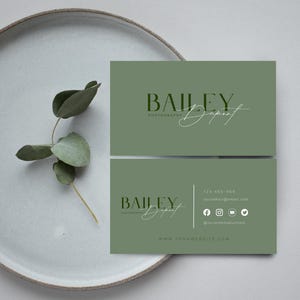 May include: Two olive green business cards with the text "BAILEY Dupont PHOTOGRAPHY" in a modern font. The bottom card includes contact information and social media icons. A sprig of greenery rests on a light grey plate.
