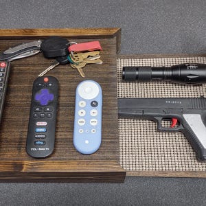 May include: A wooden tray with two compartments. The left side holds three remote controls, keys, and a pocket knife. The right side contains a flashlight and a black handgun with a red trigger.