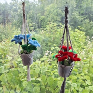 May include: Two macrame plant hangers with artificial flowers. One hanger holds blue flowers, the other holds red flowers. The white pots are suspended in front of a window with a green, leafy background.