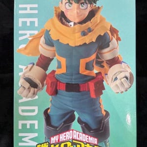 May include: A boxed action figure of a character from "My Hero Academia." The figure is of a male character with green hair, wearing a blue and orange costume. The box has the show's logo and the Bandai Namco and Banpresto logos.