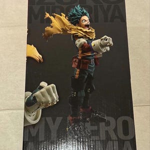 May include: A collectible figure of Izuku Midoriya from My Hero Academia, depicted in a dynamic pose with a yellow scarf, dark green suit, and large white gauntlets. The figure is displayed on a black box featuring the character's name and series title.